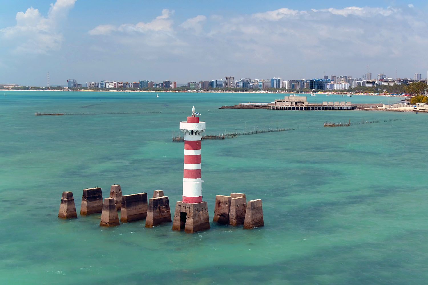 Farol sobreposto sobre pedras, água azulada e cidade ao fundo.