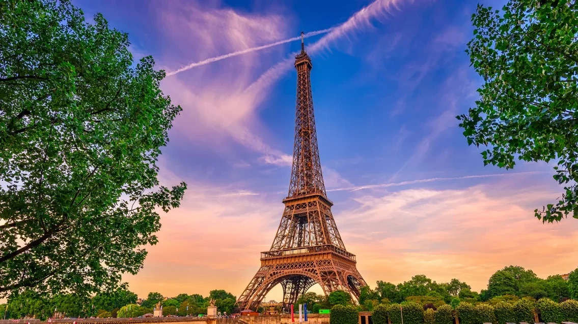 Árvores, Torre Eiffel triangular, céu colorido de azul e rosa, final de tarde.